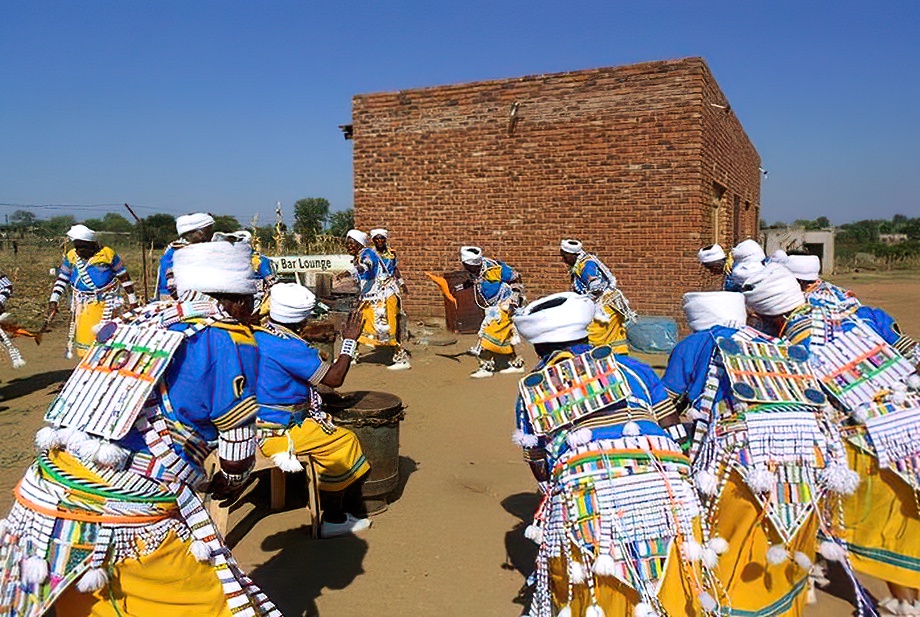 Performers in blue and yellow dress with beaded panels dancing around a drum