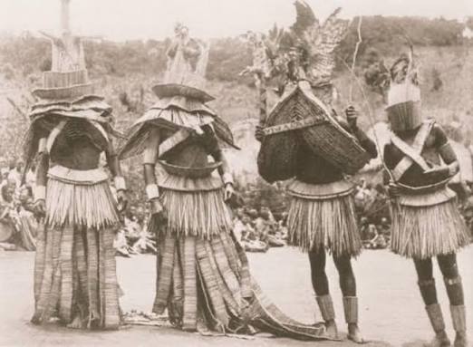 Black and white historical photograph of four people wearing elaborate ceremonial dress before a seated crowd