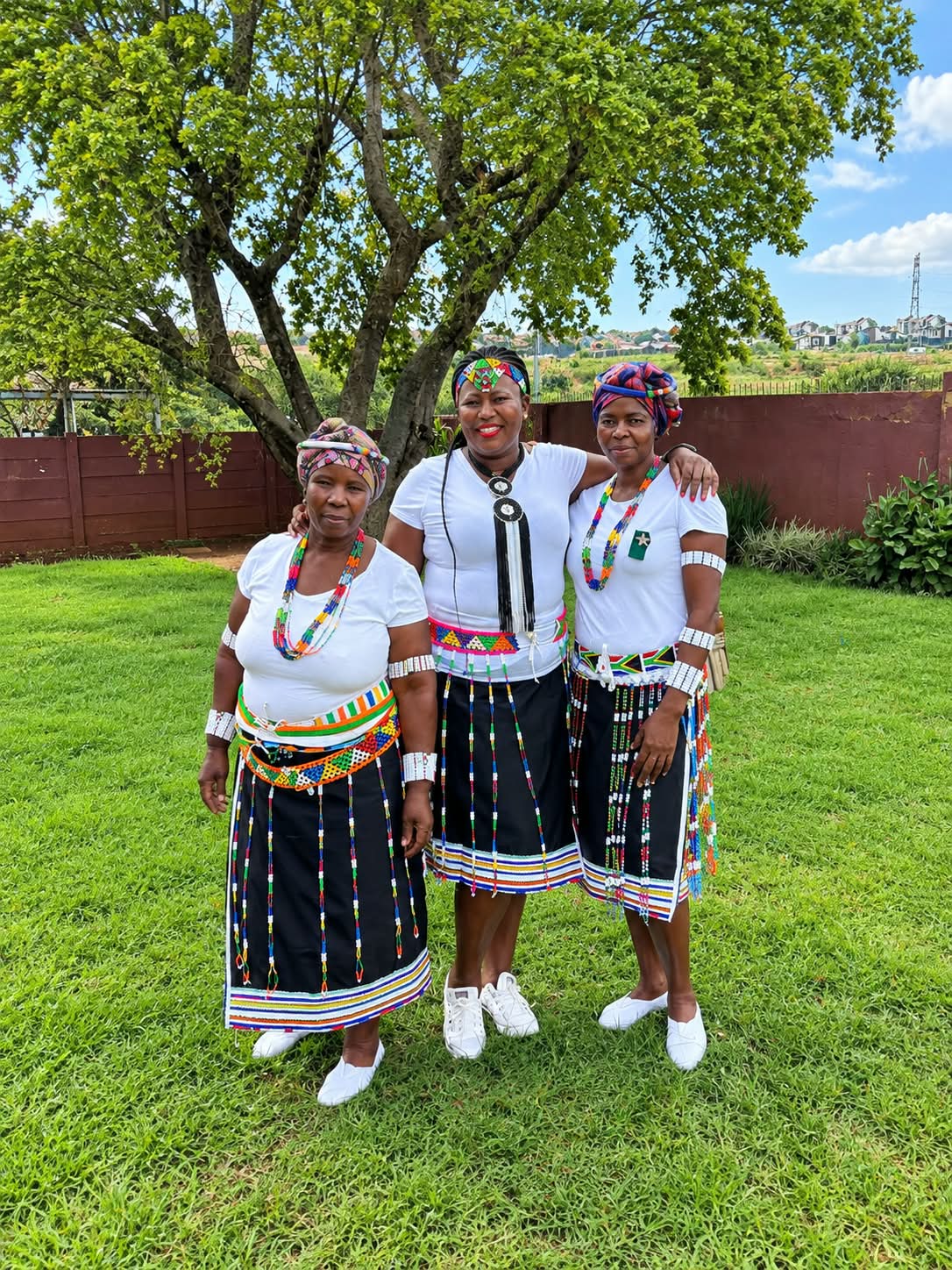 Three women outdoors wearing black skirts with colourful beadwork, white tops and patterned head wraps