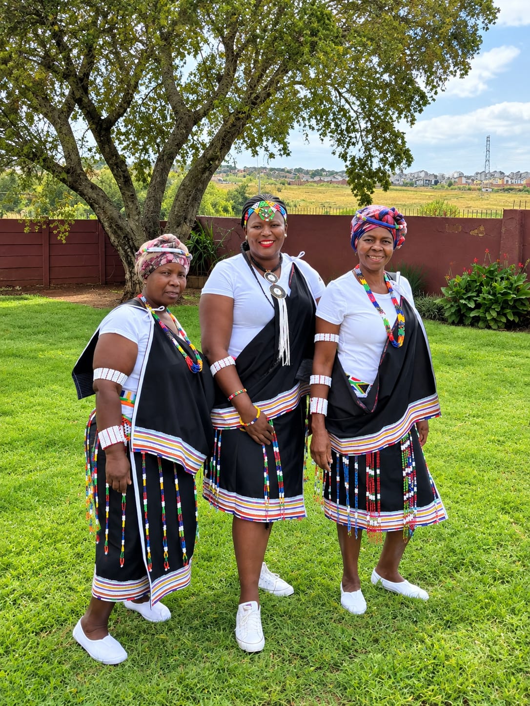 Three women standing outdoors beneath a tree in black Morina attire, white tops and colourful beadwork
