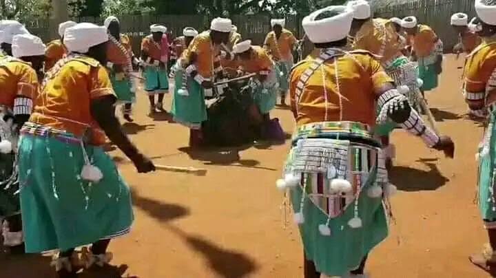 Group of dancers in orange tops, turquoise skirts and white head wraps gathered around a drum