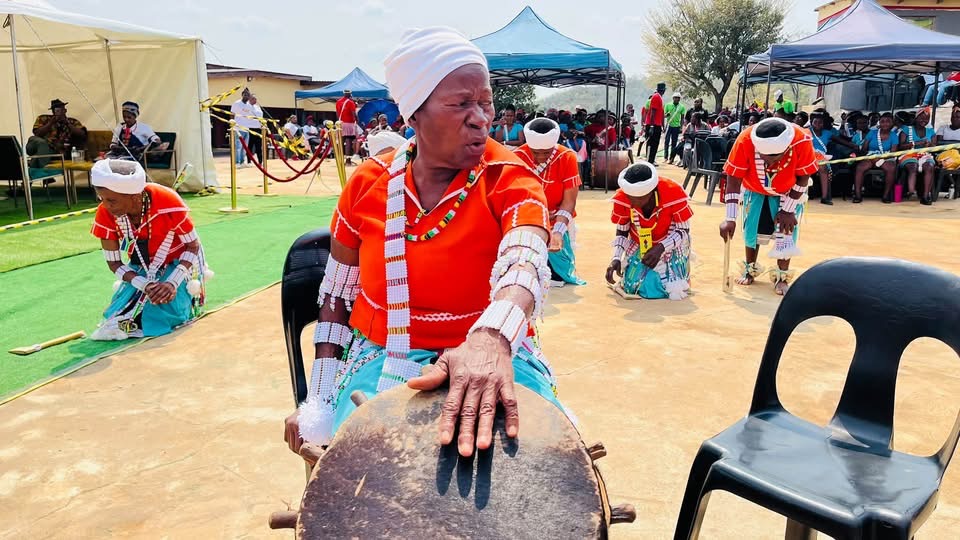 Elder playing a drum in orange and turquoise attire with dancers behind