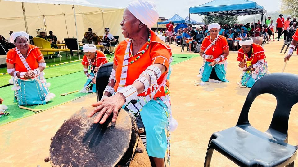 Side view of an elder at a drum and other performers kneeling behind