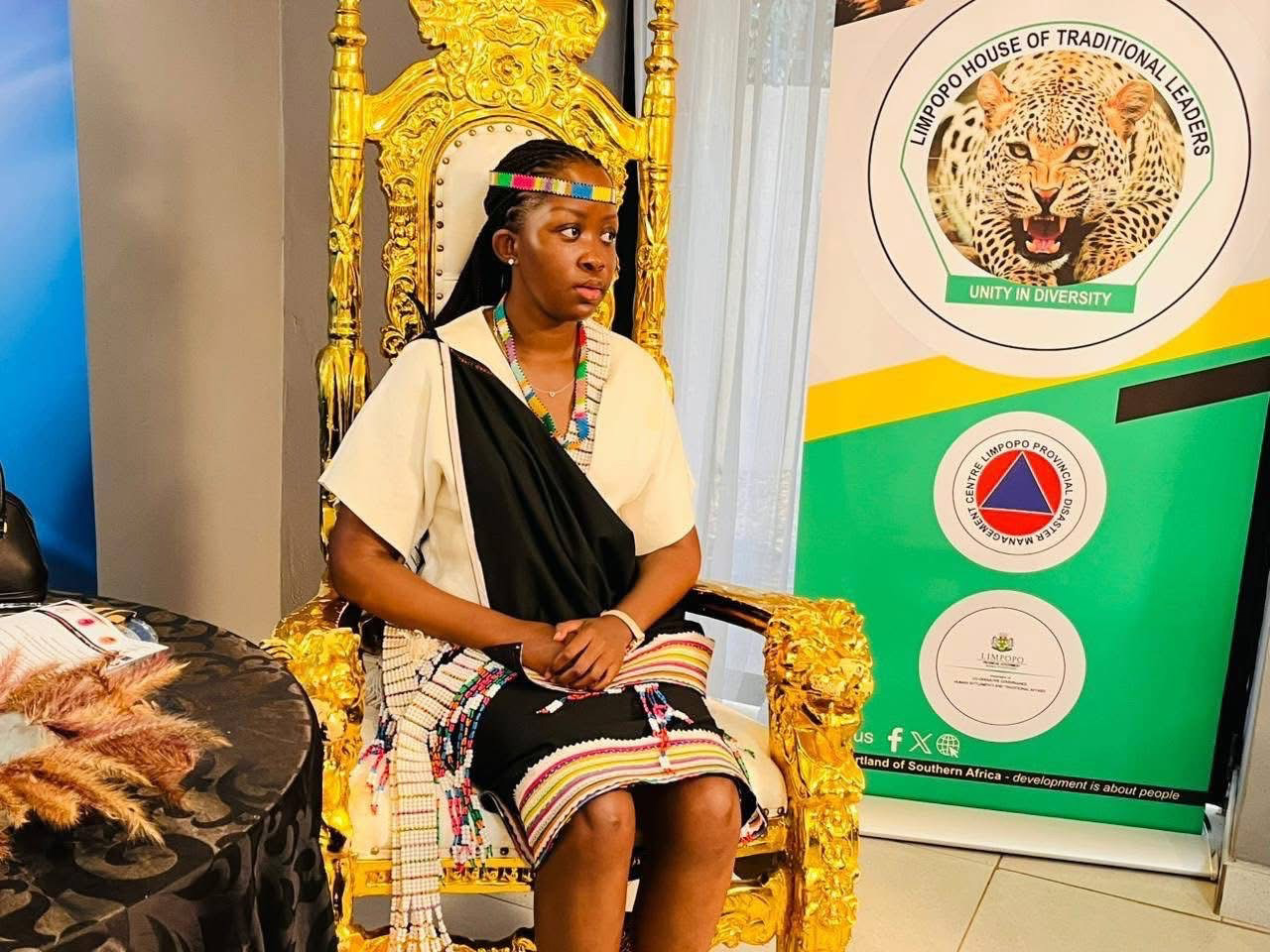 Woman seated on a gold chair wearing a black and white outfit with colorful beaded adornments
